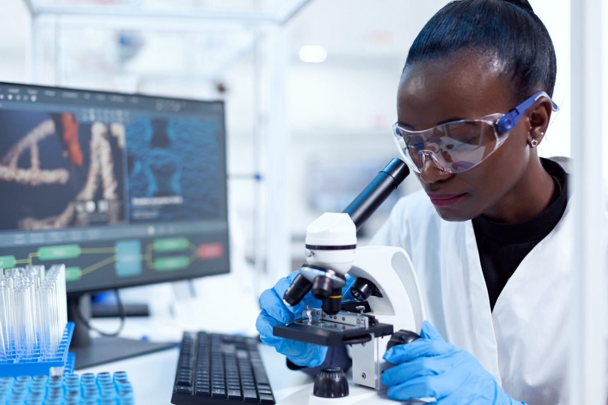 African pharmacology researcher analysing chemical substance using microscope for study.. Black healthcare scientist in biochemistry laboratory wearing sterile equipment.